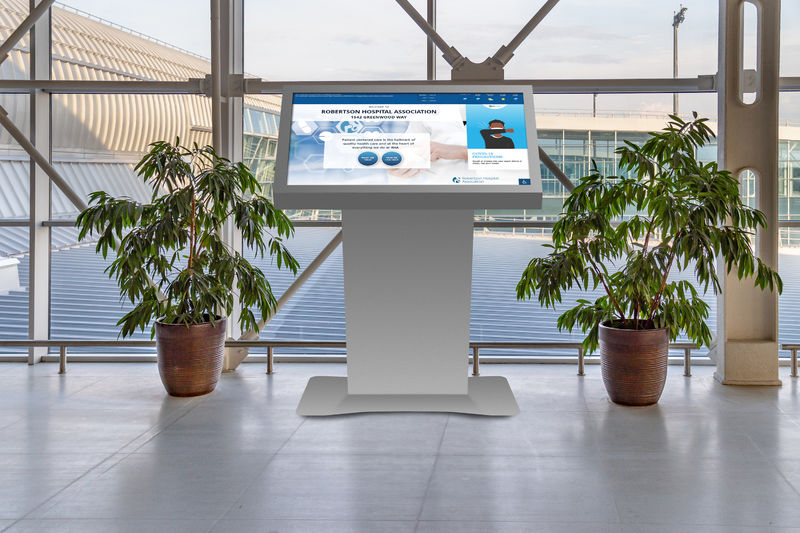 Digital interactive kiosk showing public health messaging in a hospital lobby during the daytime with potted plants on each side.