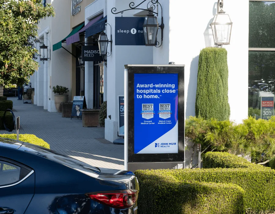 Sidewalk advertisement board for John Muir Health with blue promo, nearby storefronts and a parked car in the foreground.