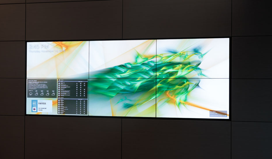 A horizontal video wall display with a green and yellow abstract digital art background and digital directory features.