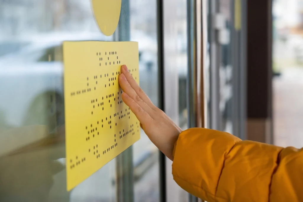 Hand from a person wearing orange puffer jacket touching a yellow braille sign on a building.