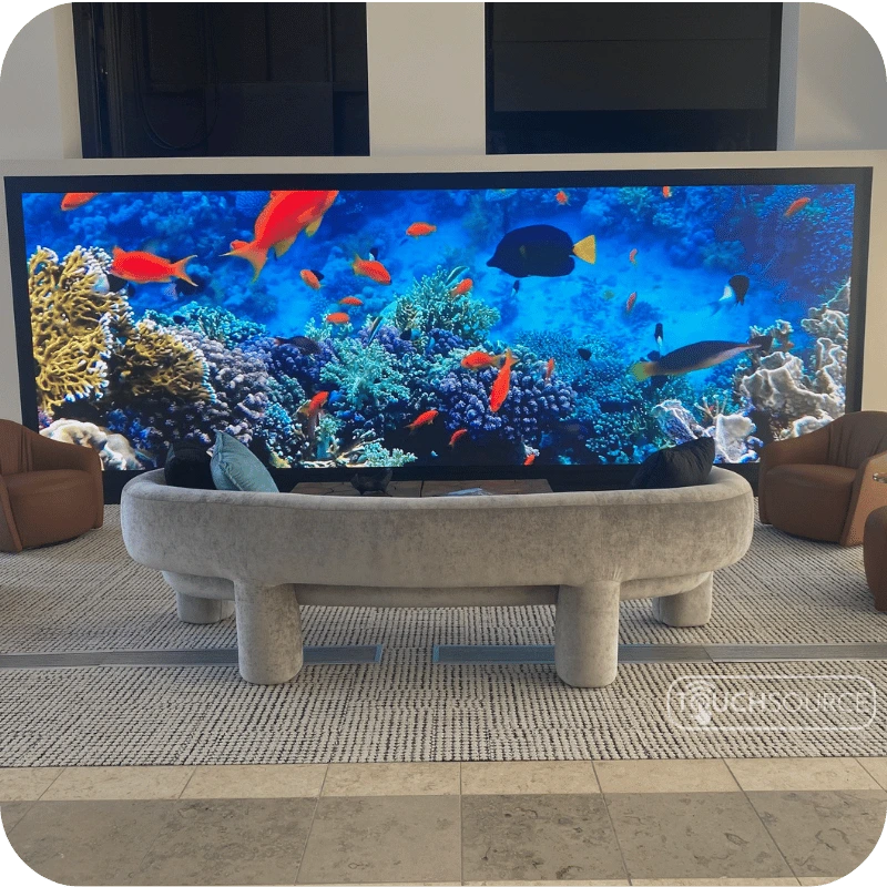 Wall-sized video screen displaying a vibrant coral reef with orange fish, in a modern lobby with a curved gray sofa.
