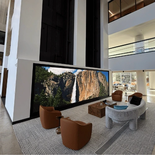 Lobby lounge with a large wall-mounted screen displaying a canyon waterfall, flanked by brown chairs and a gray curved sofa.