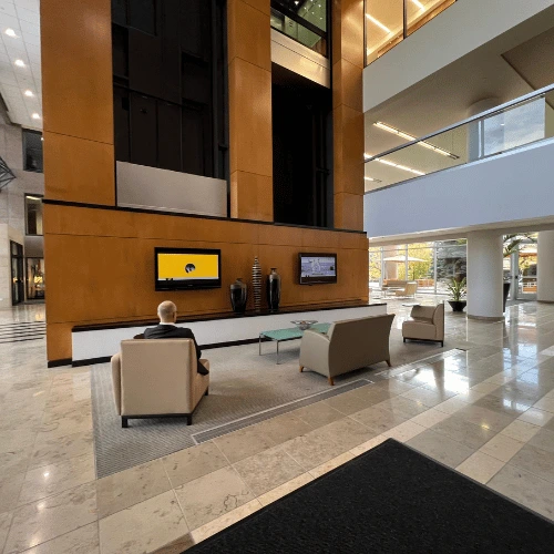 Modern hotel lobby with orange wood-paneled wall, two wall screens, and a man seated in beige chairs facing a screen.