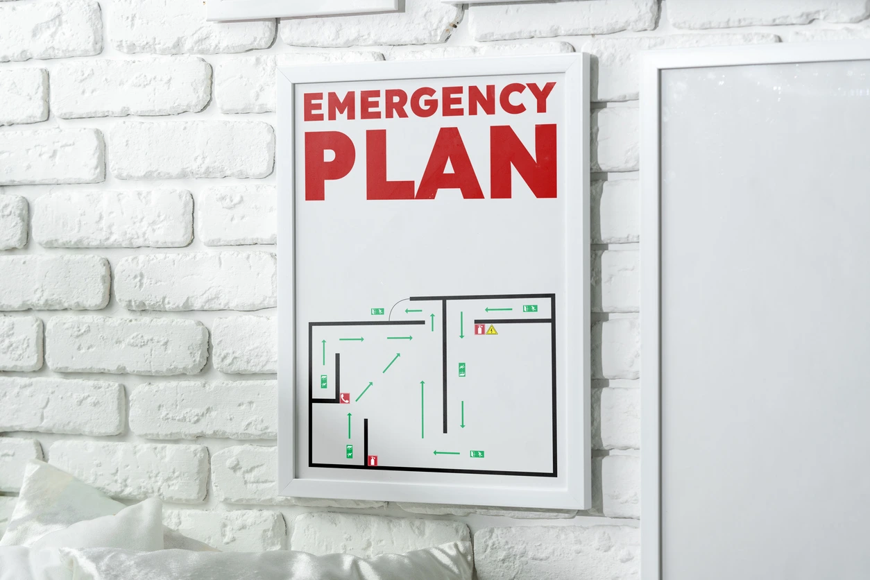 Framed white poster on a white brick wall reading 'EMERGENCY PLAN' with a simple floor plan diagram.