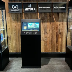 touchsource-alta-kiosk-22-inch-black-finish - TouchSource - TouchSource Self-service directory kiosk with a touchscreen showing a building image and the word DIRECTORY beneath, set against a wooden wall with brand signs above.
