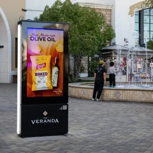 touchsource-frontier-outdoor-freestanding-kiosk-for-retail-mall - TouchSource - TouchSource Advertising kiosk in a shopping plaza featuring olive oil and Lay's Baked chips, beside a fountain and shoppers nearby