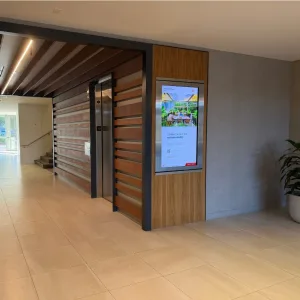 touchsource-mirada-recessed-55-inch-vertical-touchscreen-digital-directory - TouchSource - TouchSource Interior lobby featuring an elevator behind a wooden slat wall, a tall digital poster display on a wood panel, beige tile flooring, and a potted plant to the right.