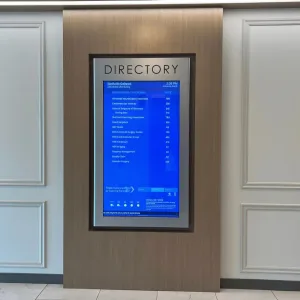 touchsource-pantella-wall-directory-portrait-vertical-orientation - TouchSource - TouchSource Digital directory screen mounted in a wood-toned stand, displaying a blue list of hospital departments and room numbers (header reads 'DIRECTORY').