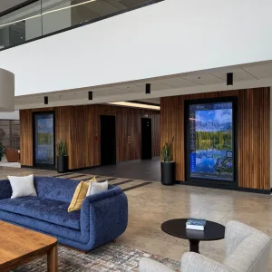 two-touchsource-recessed-mirada-85-inch-directories - TouchSource - TouchSource Modern hotel lobby seating area with a blue velvet sofa, cushions, and a round coffee table under a high ceiling. A large digital display is mounted on wood-paneled walls.